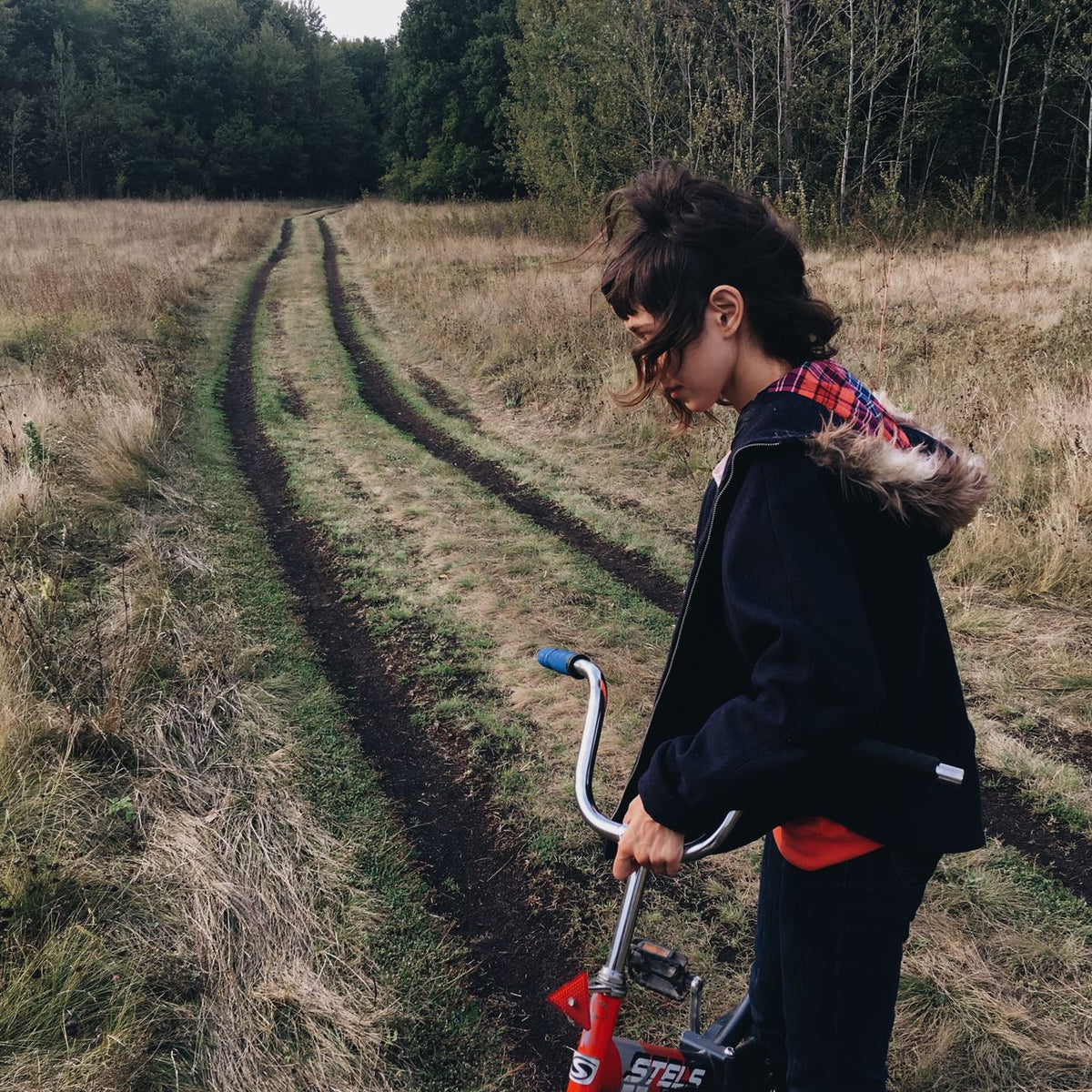 Girl standing with bicycle on a grassy country road surrounded by trees in autumn.