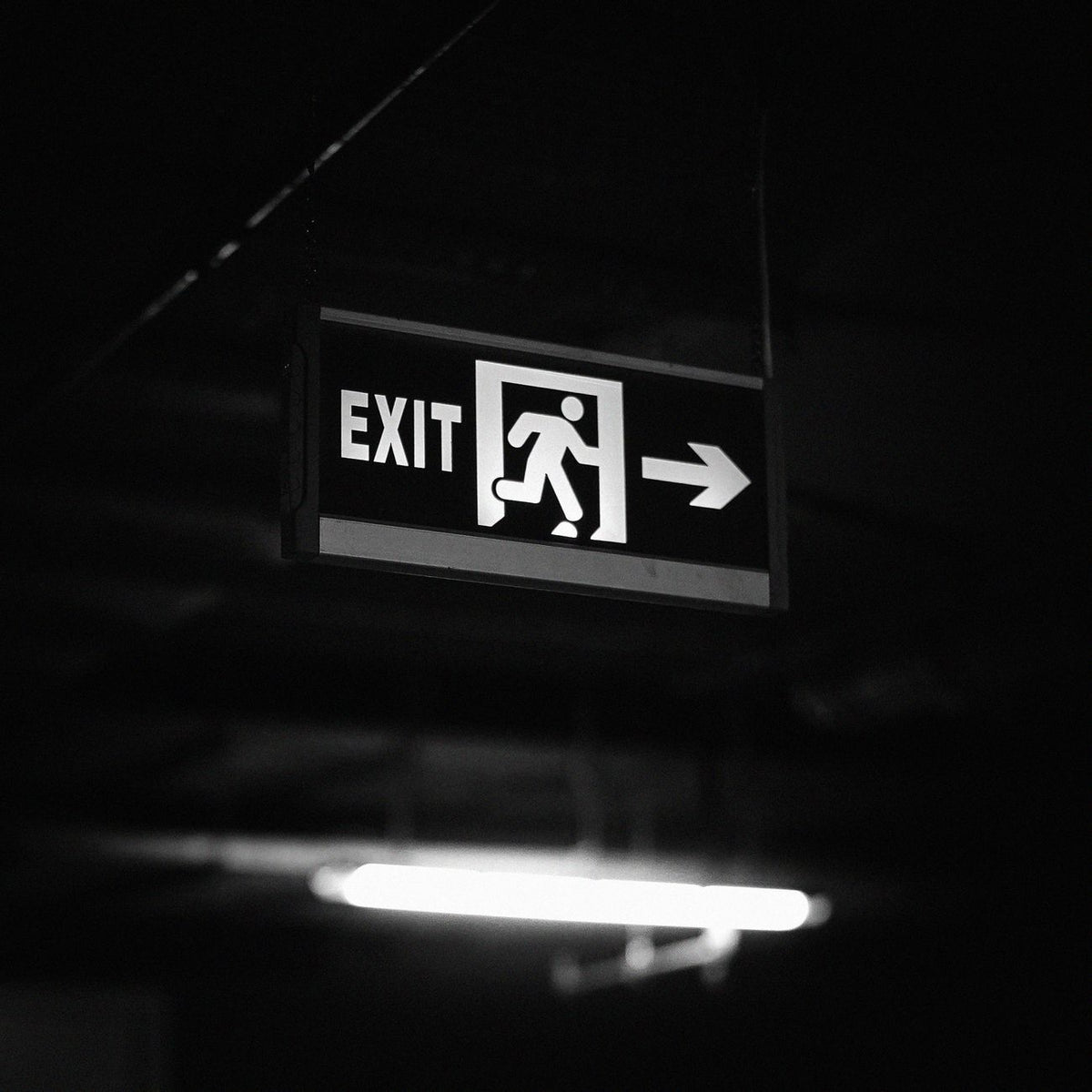 Black and white image of an exit sign illuminated in a dark setting, creating a dramatic contrast.