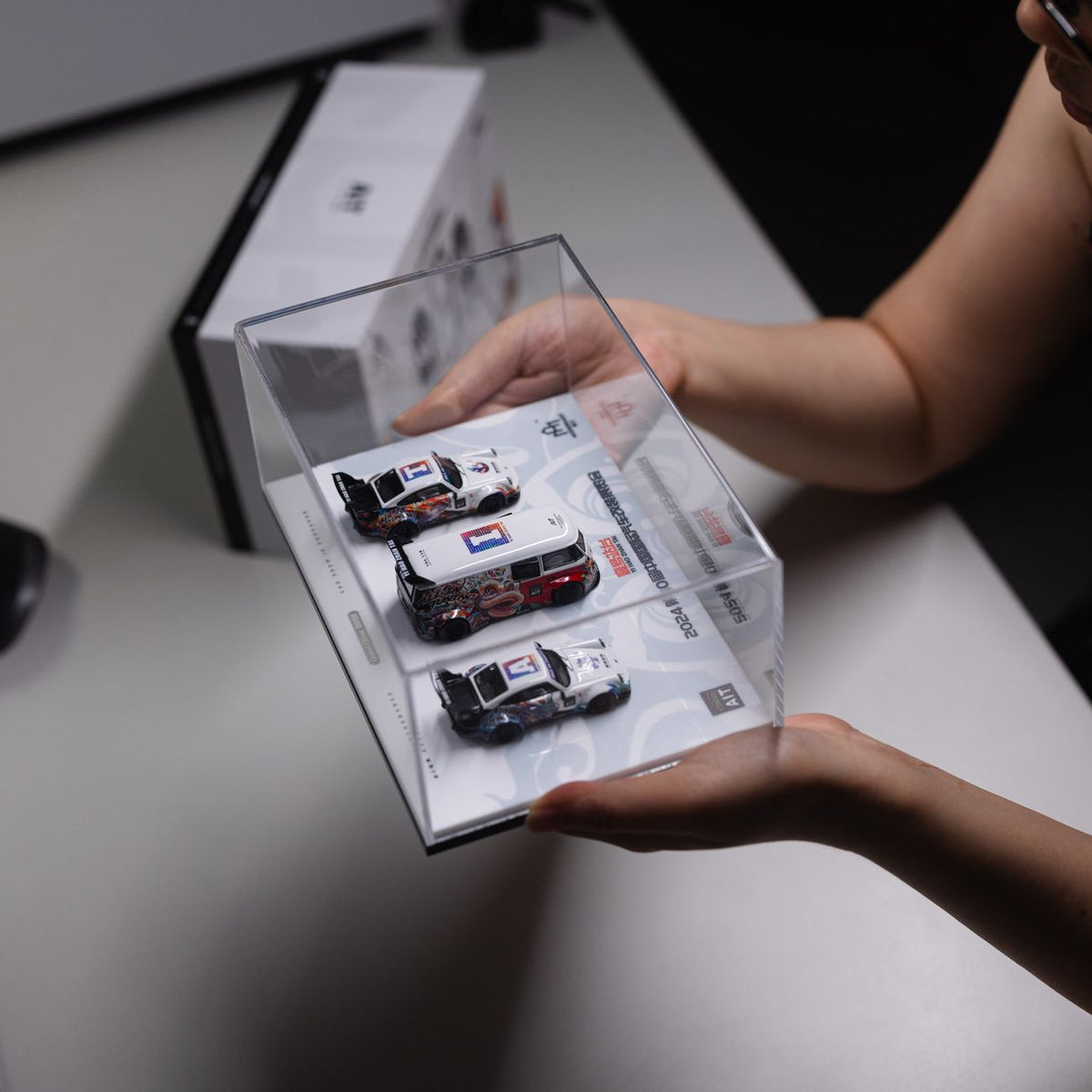 Hands holding a display case of three Porsche diecast cars showcasing minimalist design on a desktop.