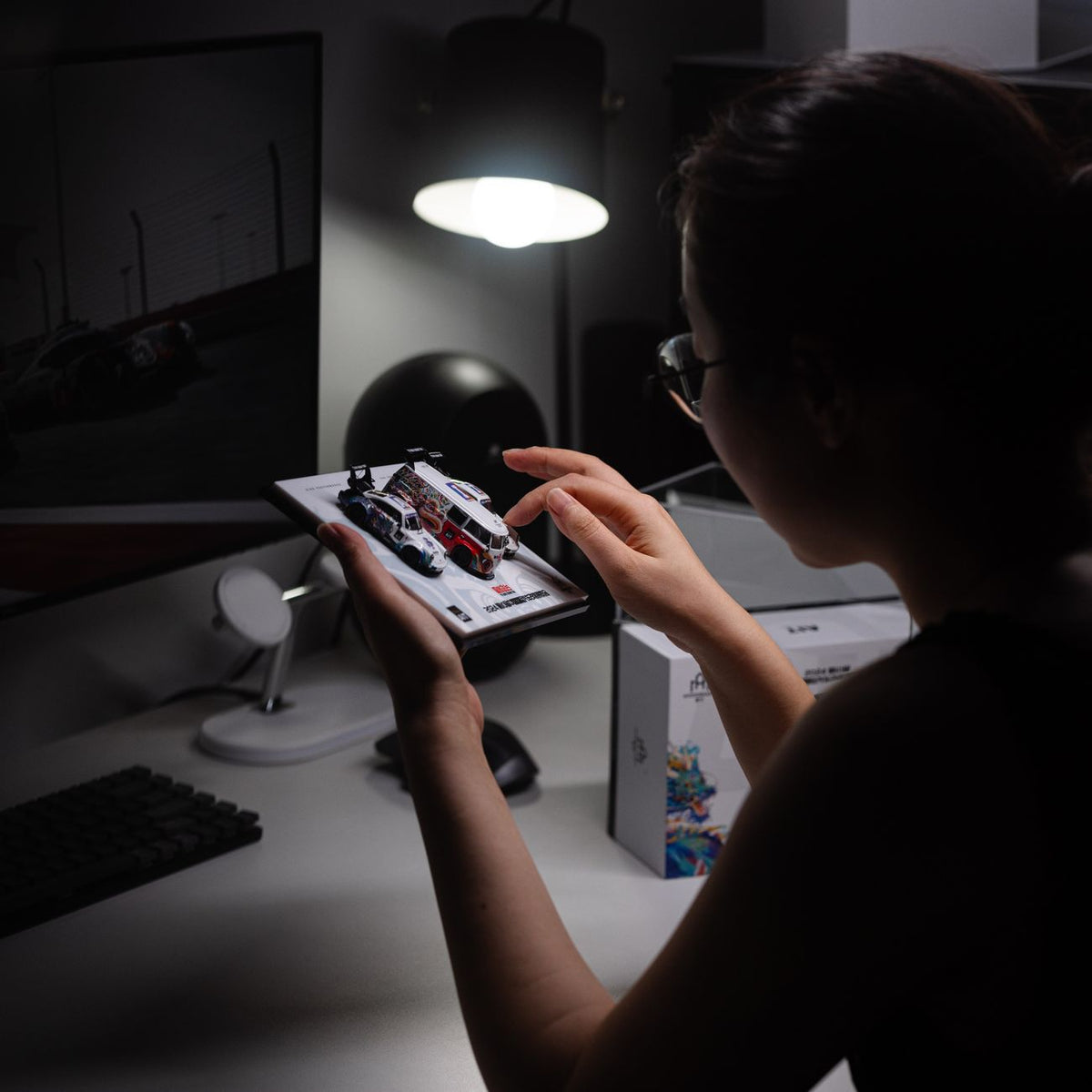 A person at a desk examines detailed diecast model cars under a lamp light.
