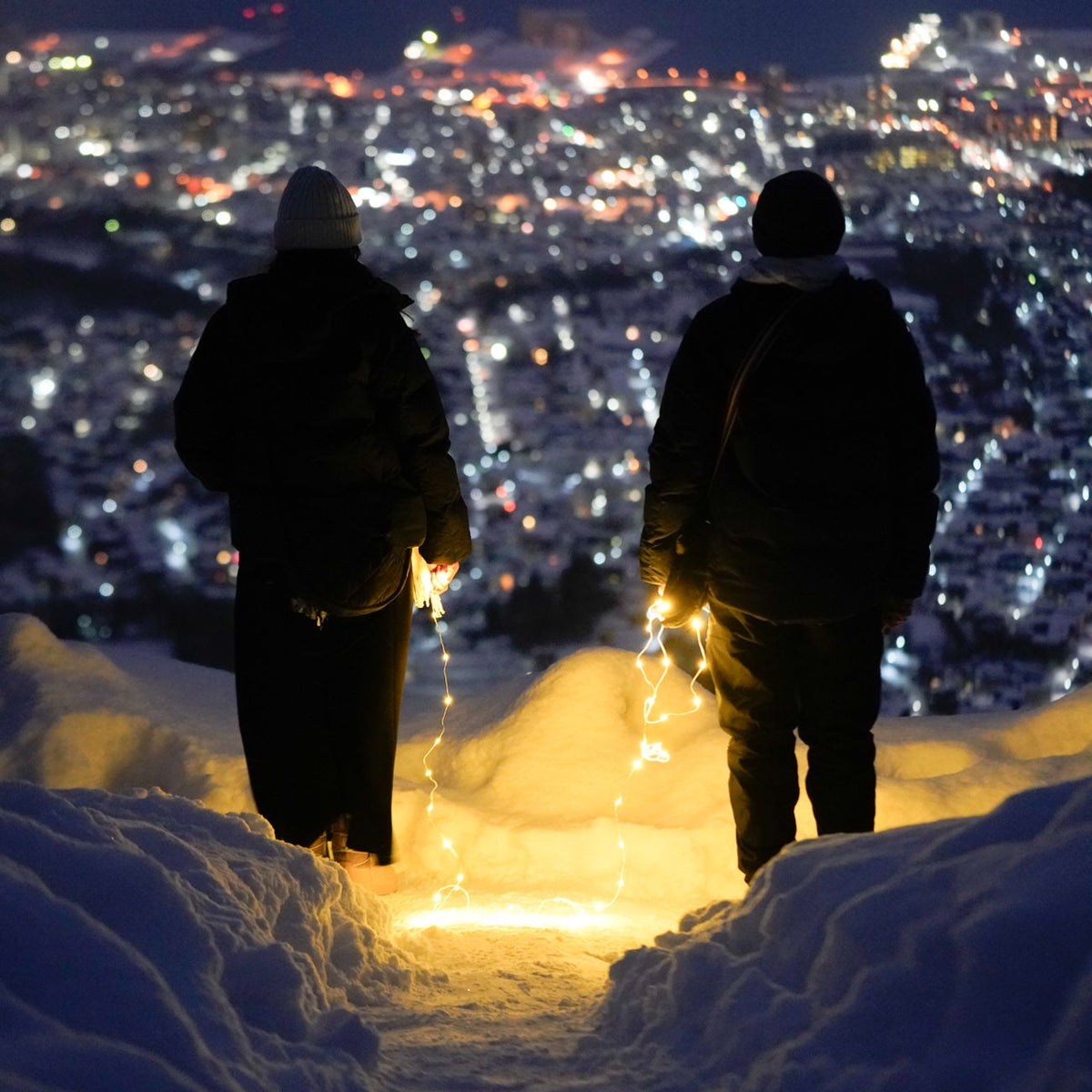 Two people holding fairy lights, enjoying a snowy cityscape at night from above.