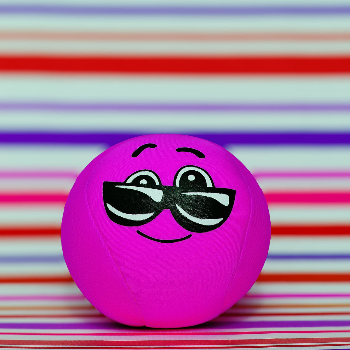 A bright pink smiley face ball wearing sunglasses on a colorful striped background.