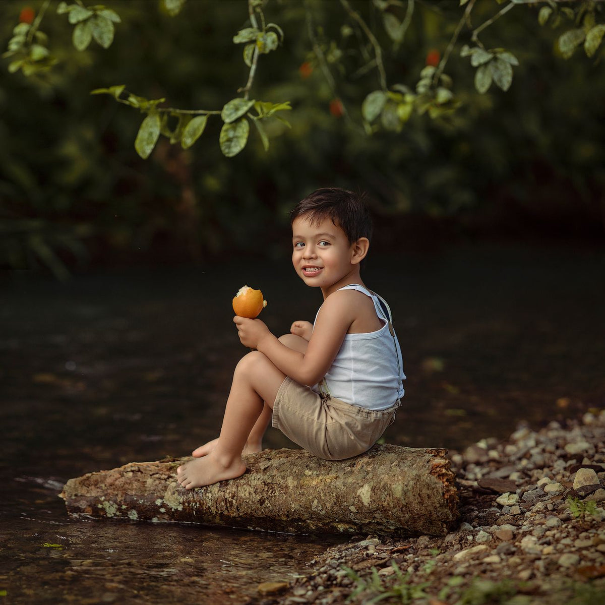 A happy child sitting on a log by a river, enjoying a mango in Tingo María, Peru.