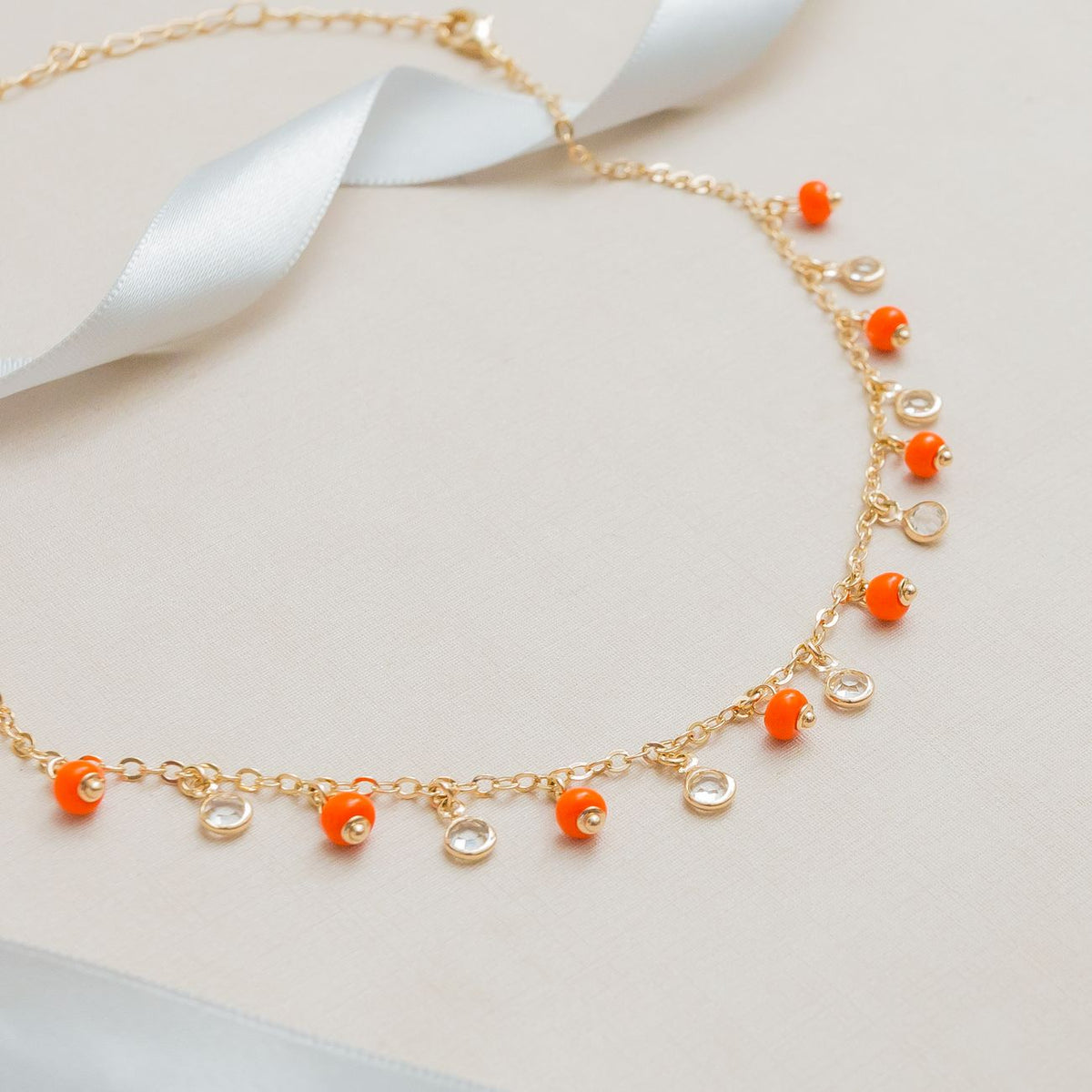 Stylish gold necklace with orange gemstones and ribbon on neutral background.