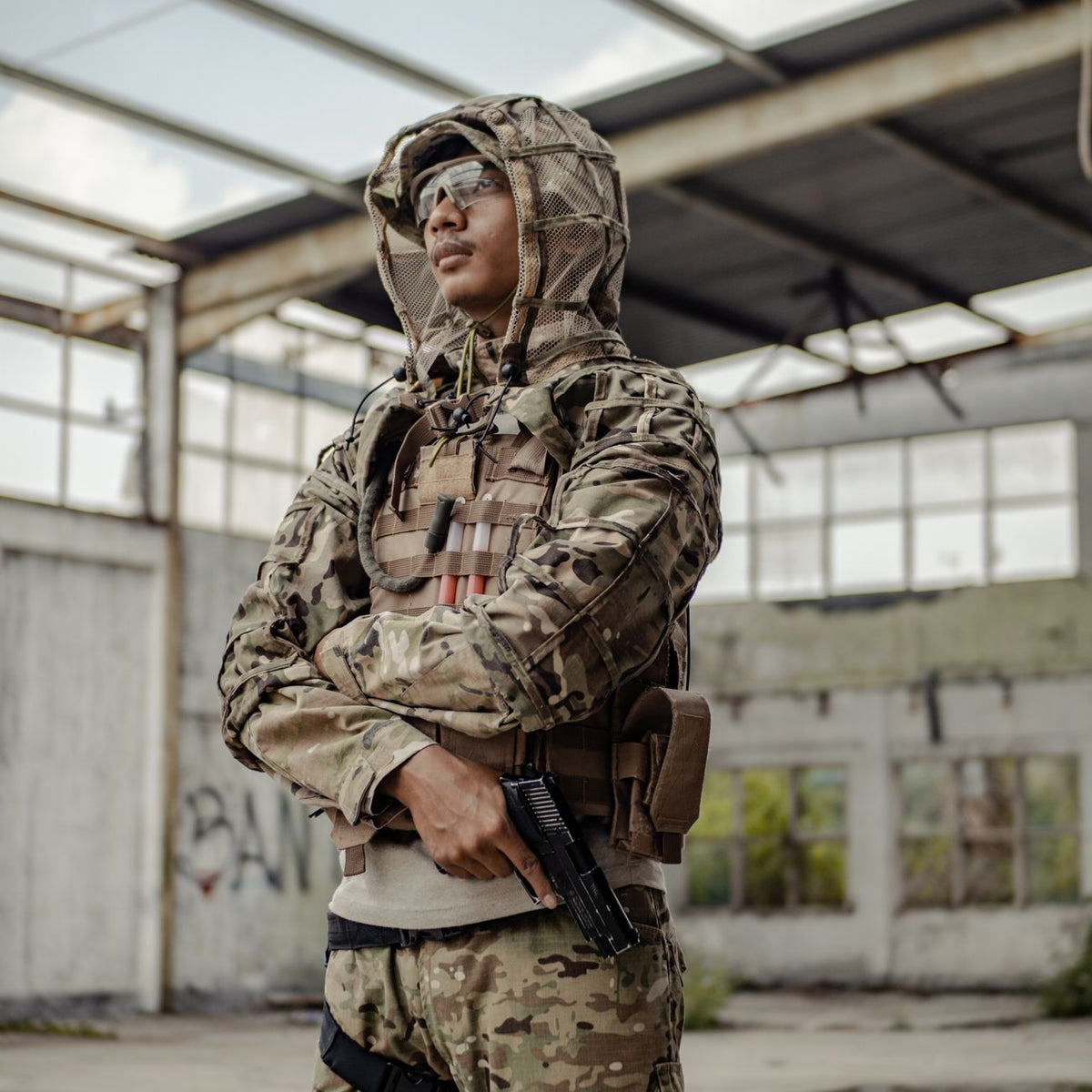 Thoughtful young ethnic soldier wearing military uniform and protective eyeglasses standing with gun and arms crossed in obsolete concrete building and looking away