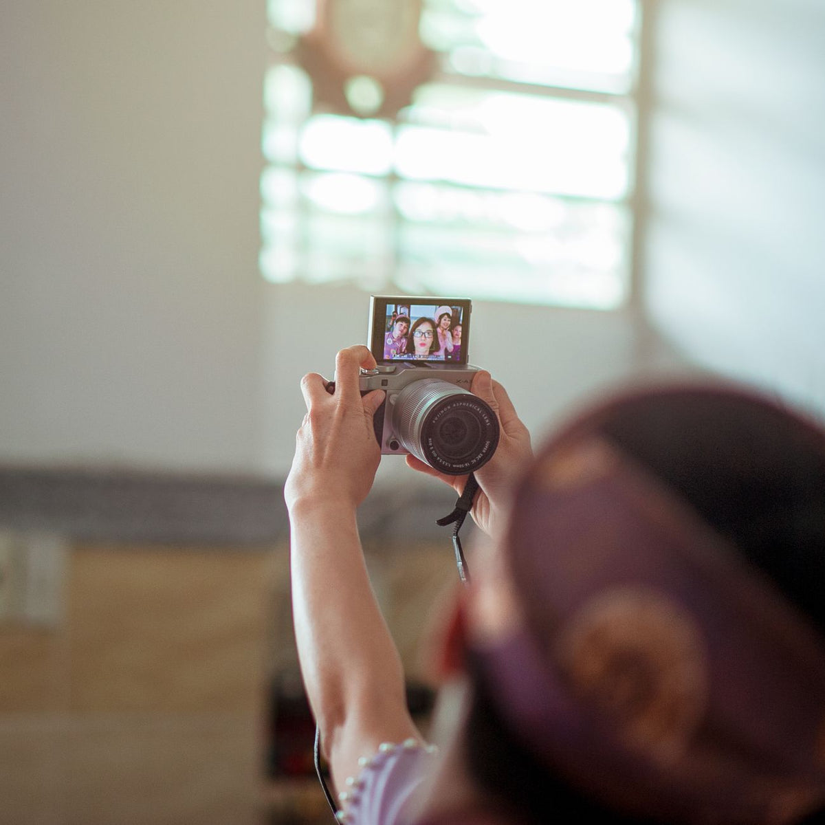 A person taking a selfie indoors using a DSLR camera, with sunlight streaming through a window.