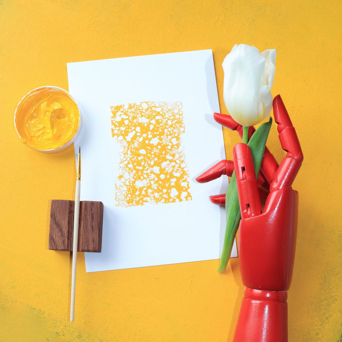 Artistic composition featuring a red robotic hand holding a tulip with paint supplies on a yellow background.